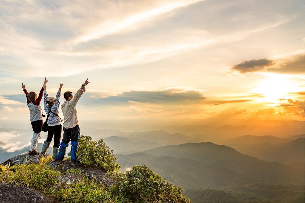 Three hikers with their arms raised in excitement at the top of a mountain with a beautiful view in the distance.