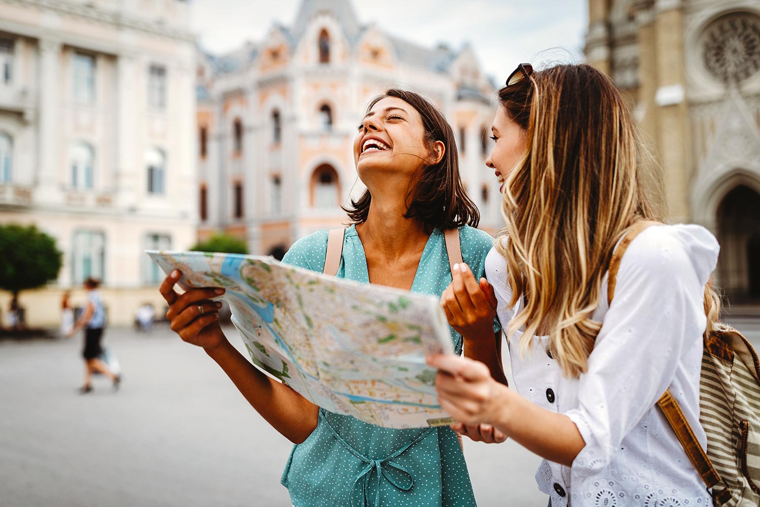 Two young female travelers holding a map and laughing in a European city square.