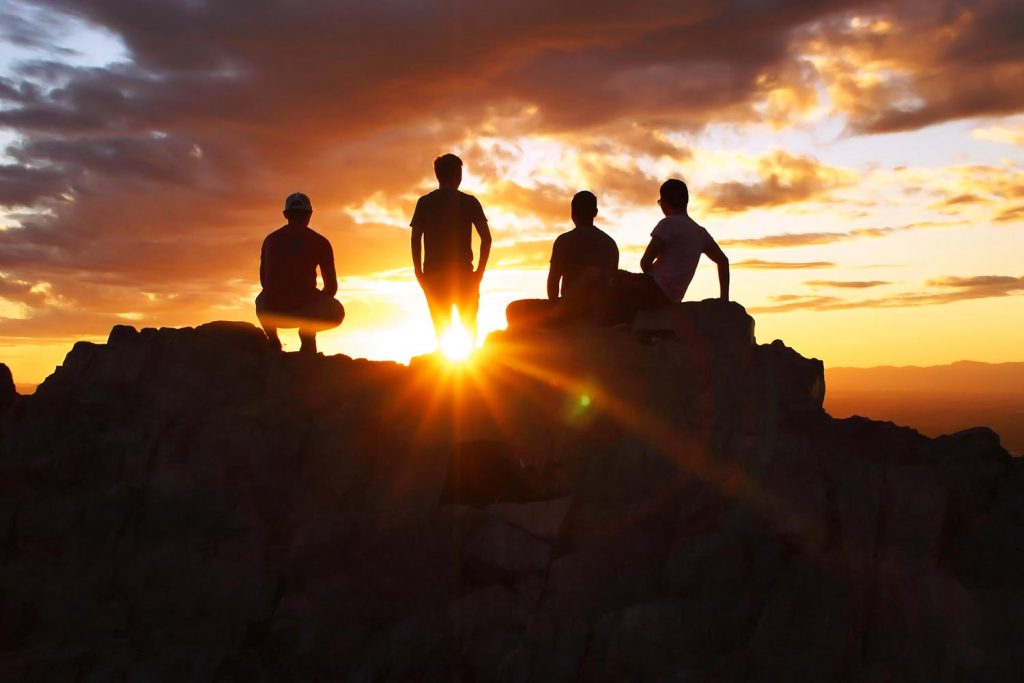 Four men silhouetted on the top of a mountain with a stunning sunset behind them. 